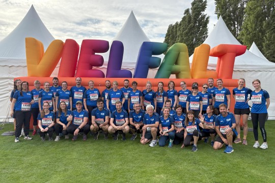 Auf dem Bild ist eine große Gruppe von Menschen in blauen Sporttrikots zu sehen. Sie stehen vor einem großen bunten Vielfalt-Schriftzug