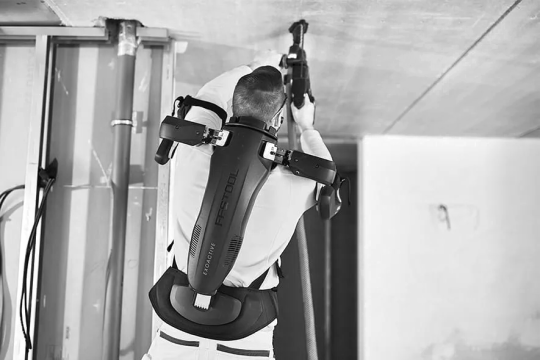 Auf dem Bild ist eine Person zu sehen, die in der Decke bohrt und dabei ein Exoskelett auf dem Rücken trägt.