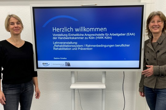 Das Bild zeigt Sabine Gnielka links und Professorin Mathilde Niehaus rechts neben einem Bildschirm. Auf dem Bildschirm steht: Herzlich wilkommen. Vorstellung Einheitliche Ansprechstelle für Arbeitgeber (EAA) der Handwerkskammer zu Köln (HWK Köln).