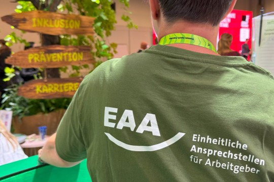 Auf dem Bild ist eine EAA Fachberatenden von hinten zu sehen. Auf ihr T-Shirt ist das Logo der EAA gedruckt und sie steht vor Wegweisern beschriftet mit Inklusion, Prävention und Karriere.