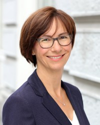 Potrait von Frau Handke: Sie hat kurze braune Haare und eine dunkle Brille. Sie trägt einen blauen Blazer.