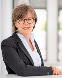 Potrait von Frau Schartz: Sie hat kurze graue Haare, träg eine braune Brille und trägt eine weiße Bluse mit einem grauen Jacket.